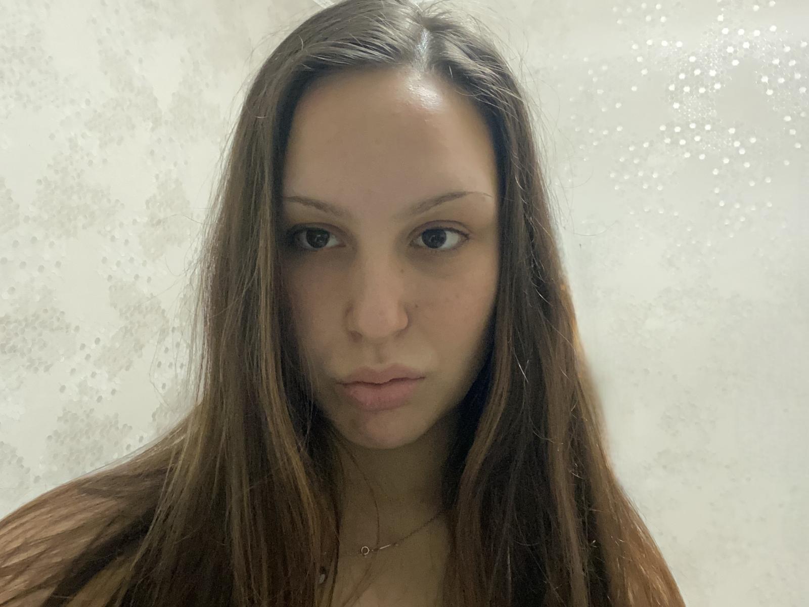 TishaEgge ha lunghi capelli castani, pelle chiara e labbra piene. Guarda direttamente l'obiettivo con un'espressione neutra. Si vede una piccola collana..