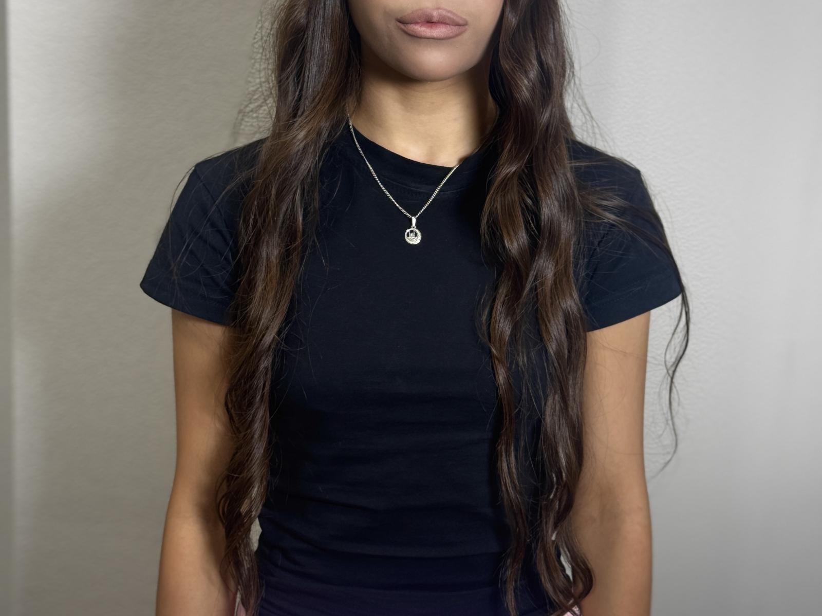 AliceVljeric with long, wavy brown hair, wearing a black shirt, and a silver necklace with a circular pendant, showing her tits and a subtle ass.
