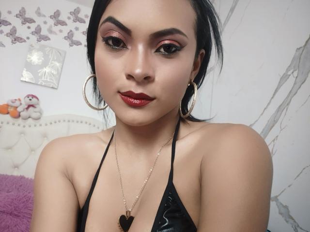 Kalie Bennett mit glänzenden roten Lippen, schwarzem Eyeliner, Reifenohrringen, einer Goldkette und einem schwarzen Halstopp, der ihre Brüste zeigt, in einem Selfie mit einem selbstbewussten, sexy Blick..