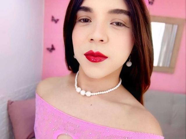 AndrewTaylor69 tiene lápiz labial rojo, cabello castaño oscuro, una blusa rosa con un hombro descubierto, un collar de perlas blancas y aretes, con paredes rosas y pegatinas de mariposas en el fondo..