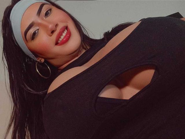 CristallWrightTs a de grandes seins, elle porte un haut noir avec une découpe, du rouge à lèvres, des boucles d'oreilles pendantes et un bandeau bleu, elle sourit à l'appareil photo et a les cheveux foncés..