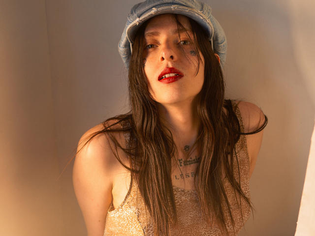 Sasha, con i suoi lunghi capelli castani, il rossetto rosso, indossando un berretto blu e una maglia di pizzo, ha tatuaggi sul petto e sul collo, e guarda seducentemente nella fotocamera..