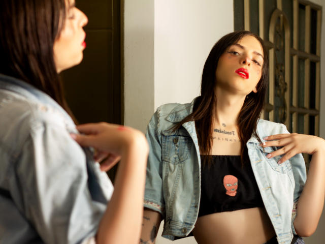 Sasha, con rossetto rosso, top nero che lascia intravedere piccoli seni, giacca di jeans e tatuaggio, si guarda allo specchio. Il suo riflesso mostra la sua mano con le unghie dipinte..