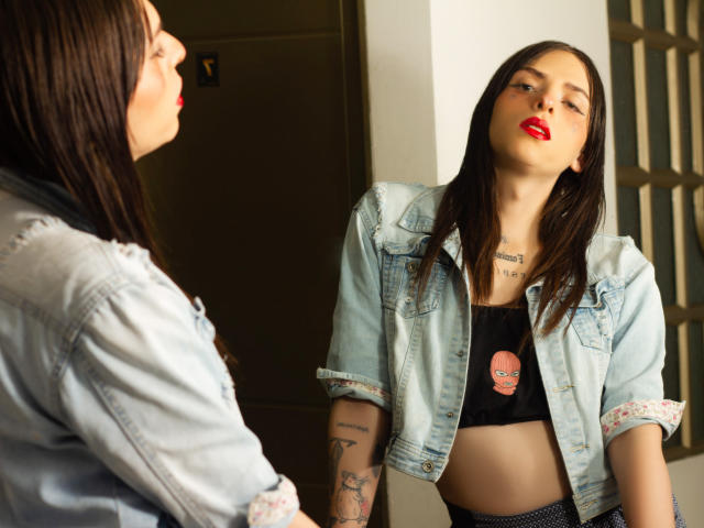 Sasha, con le labbra rosse, una maglietta nera che mette in mostra un seno piccolo, una giacca di jeans e tatuaggi, è in piedi di fronte a un'altra donna con i capelli castani e una giacca di jeans. Sembra sicura di sé, con lo sguardo leggermente rivolto verso l'alto..