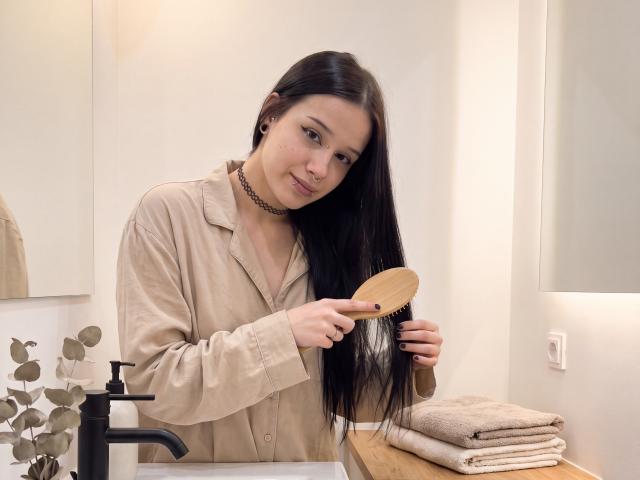 LadyKezele mit langem, schwarzem Haar, einem beigen Hemd und einer Halskette, hält einen hölzernen Spiegel in einem Badezimmer, zeigt auf ihr Spiegelbild, trägt schwarze Ohrringe, ein cremefarbenes Handtuch liegt auf einer hölzernen Ablage..