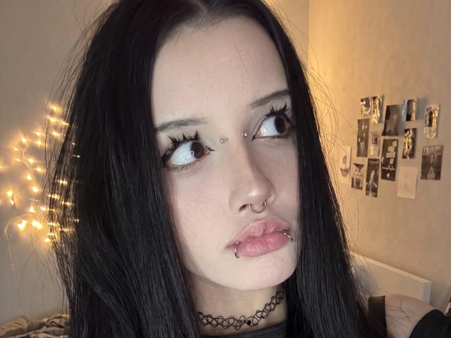 LadyKezele met zwart haar, bleke huid en meerdere piercings in haar gezicht (neus, lip en boven de wenkbrauw). Ze draagt een zwarte choker en kijkt naar de zijkant, met lange wimpers..