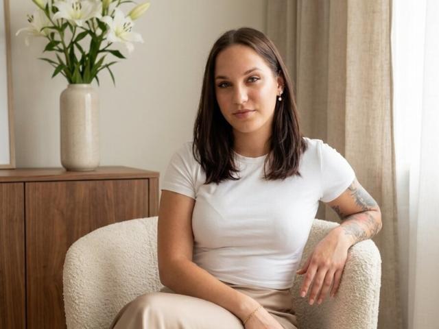 WinnieFalso con capelli castani, maglietta bianca, seno piccolo, braccio tatuato, seduta su una sedia beige, fiori bianchi su un mobile in legno, tenda color crema, sorriso leggero, posa rilassata..