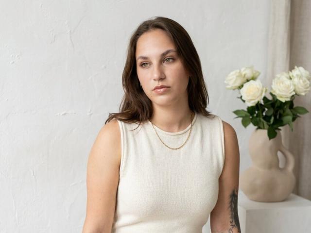 WinnieFalso, com cabelo castanho, vestindo uma blusa branca sem mangas, um colar de ouro e uma tatuagem no braço direito, sentada com rosas brancas em um vaso bege..