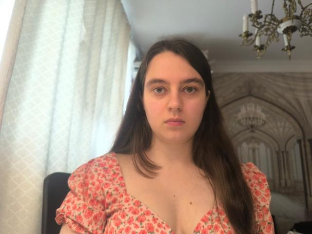 CheryllBlurton with long brown hair, pink floral dress, and small tits, standing in front of white curtains, looking directly at the camera with a neutral expression.