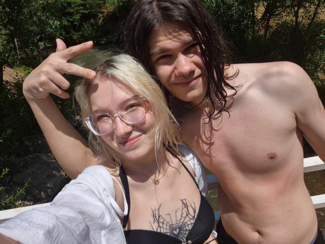 EllisSamain with blonde hair, glasses, and black bikini top, showing a peace sign, next to a shirtless EllisSamain with wet brown hair and small nipples, smiling outdoors.