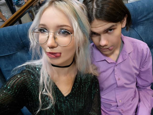 EllisSamain mit blonden Haaren, Brille und einem grünen, mit Pailletten besetzten Oberteil, Zunge heraus, schwarzer Choker, neben EllisSamain in einem lila Hemd, braunen Haaren, die nach oben schaut. Auf einem blauen Sofa..