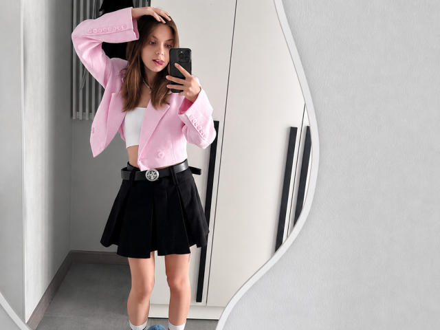 Brigette Laurie, com seios médios, vestindo uma jaqueta rosa, cropped branco, saia preta e cinto, tirando uma selfie no espelho, com a mão no cabelo, em um quarto moderno..