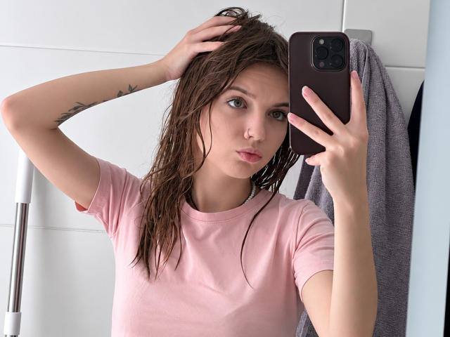 Brigette Laurie, com cabelo castanho molhado, camisa rosa, segurando um telefone, braço levantado, tatuagem visível, piercing no nariz, olhando para o espelho, parede azul clara, toalha cinza, superfície refletora..