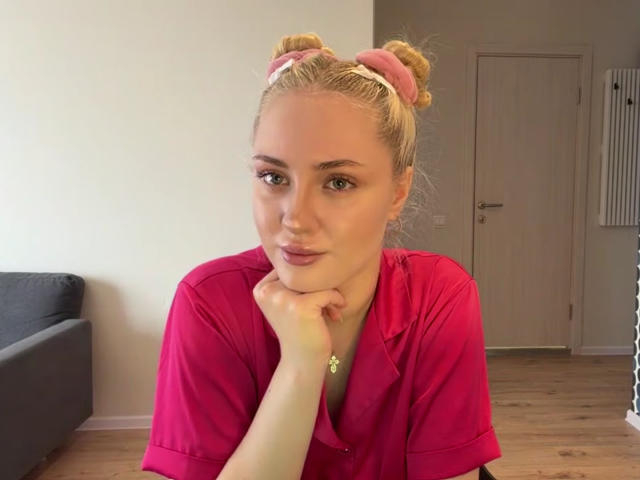 NancyBijou, con i capelli biondi raccolti in due chignon rosa, indossa una camicia rosa, appoggia il mento sulla mano, sorride dolcemente, ha gli occhi verdi e la pelle chiara, il pavimento è di legno, sullo sfondo si vede una porta..