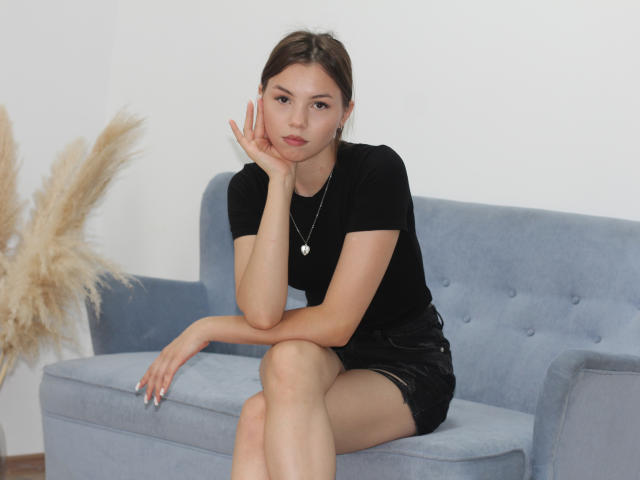 AgataRyili sitzt auf einem blauen Sofa, trägt ein schwarzes T-Shirt und schwarze Shorts, hat eine Hand am Kinn, blickt in die Kamera, mit einem nachdenklichen Ausdruck, trägt eine Halskette und hat einen prallen Hintern..