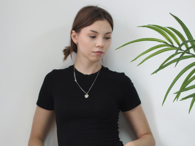 AgataRyili, vêtue d'un t-shirt noir, a de petites seins et un collier fin avec un pendentif en forme de cœur. Elle se tient debout, avec une expression pensive, à côté d'une plante verte..