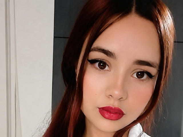 LunaSophiee heeft glanzend bruin haar, grote bruine ogen met dikke zwarte eyeliner, en rode lippenstift. Haar gezicht is glad met een lichte huidskleur en een schattige, serieuze uitdrukking..
