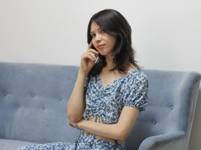 LisaSeili, mit mittelgroßer Brust, trägt ein blaues, geblümtes Kleid und sitzt auf einem grauen Sofa. Sie hält ihr Telefon ans Ohr, hat ein leichtes Lächeln und dunkles, welliges Haar..