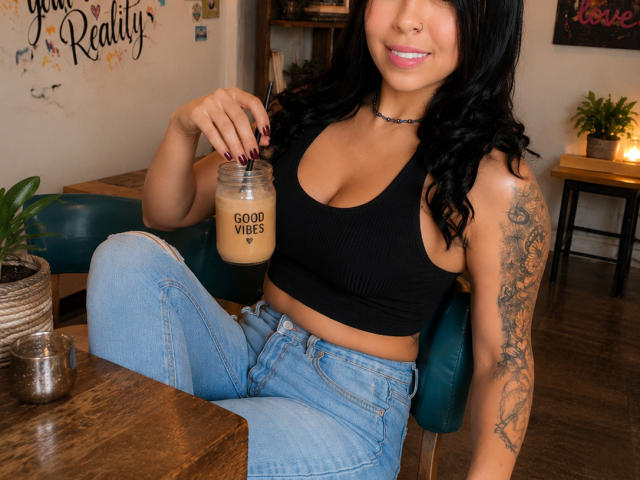 NenaBlack with black hair, tan skin, and tattoos, sips "Good Moods" latte, wearing a black ribbed tank and blue jeans, sitting in a cozy cafe.