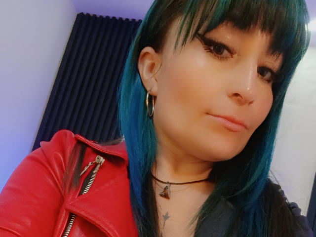 AliceSweetLady, con cabello verde azulado, vistiendo una chaqueta de cuero roja, un collar negro y aros grandes, sonriendo ligeramente, mostrando un pequeño piercing en la nariz y un tatuaje en el pecho..