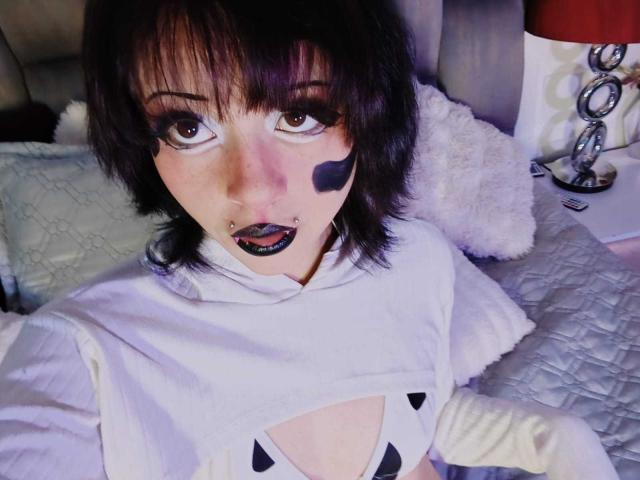ViolettClay with dark purple eyes, black lipstick, black facial paint, black nose ring, white shirt, and short black hair, lying on a bed with a white pillow.
