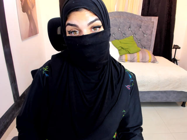Aniisa Ali, con un hijab negro, una máscara negra que cubre su nariz y boca, un hijab bordado en verde y azul, sentada en un dormitorio, con un cojín verde, una cama gris y cortinas amarillas en el fondo..