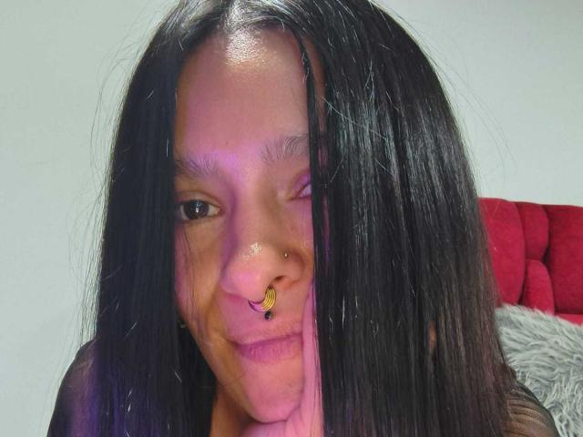 AmelieBlake has long black hair, a nose ring with a small yellow butterfly, and is winking with a slight smile. Her skin is tan and she's looking cute.