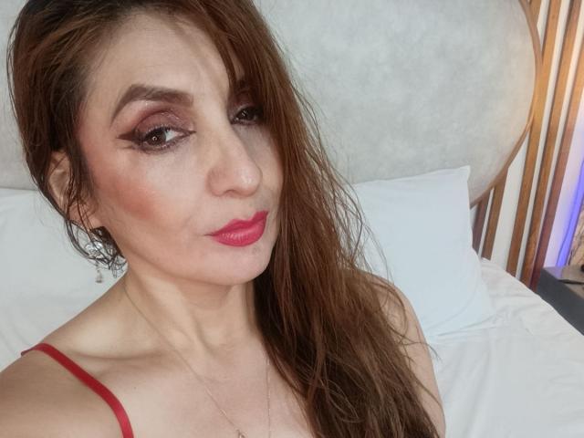 ElenaHart heeft nat bruin haar, rode lippenstift en smoky oogmake-up, met haar borsten licht zichtbaar onder een rode top. Ze kijkt met een subtiele glimlach in de camera..