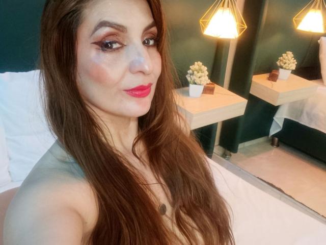Elena Hart, met lang bruin haar, rode lippenstift en veel make-up, laat haar borsten zien op een selfie, zonder top, in een helder verlichte kamer met twee hangende lampen..