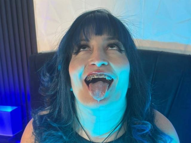 AliceSweetLady with blue hair, tongue out, drooling, metal teeth, looking up, blue light on face, black sleeveless top, black hair, and a black background.