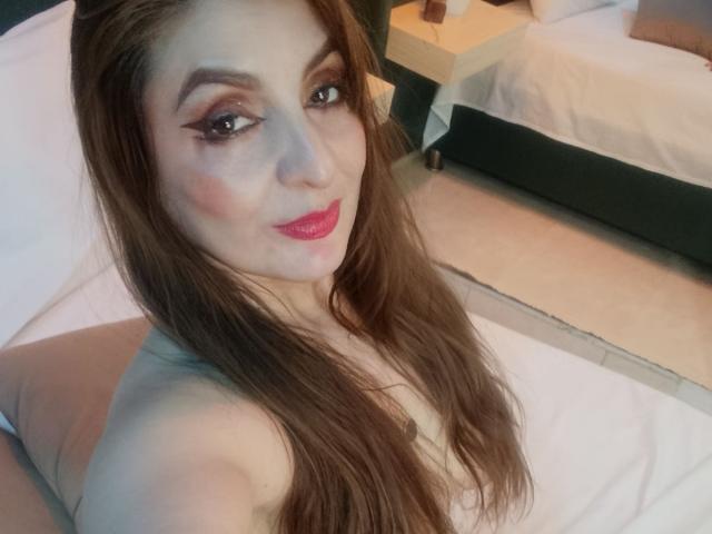 Elena Hart, con cabello castaño largo, lápiz labial rosa y delineador de ojos, sin ropa, mostrando sus senos, acostada en una cama, mirando a la cámara, sonriendo, en un dormitorio..