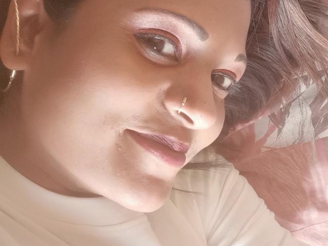 El rostro de IndianLusty, ligeramente inclinado, muestra un pequeño piercing en la nariz y sombra de ojos rosada. Ojos marrón claro, una sonrisa sutil, labios rosados y piel marrón claro. Una luz suave resalta sus rasgos..