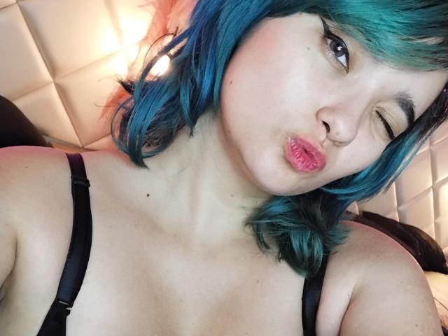 Naommy Carter, con cabello azul verdoso, guiñando un ojo, labios rosados apretados, vestido con tirantes negros, piel clara, una pequeña peca visible en el cuello, con la cabeza apoyada en un almohadón blanco..
