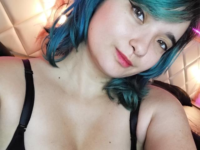 Naommy Carter, con cabello azul turquesa, un sostén negro, labios rosados y piel clara, sonriendo a la cámara, mostrando sus senos y un rostro atractivo. Iluminación suave en el fondo..