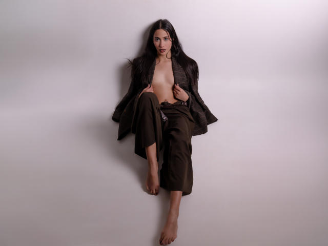 JennTylor with long black hair, naked except for an open black jacket and green pants, barefoot, legs crossed, holding jacket, small tits, confident expression, studio lighting.