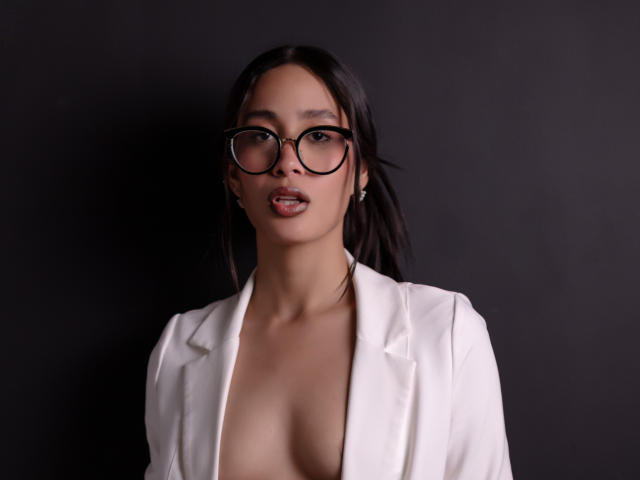 JennTylor with black-rimmed glasses, white open jacket, and exposed boobs, standing against dark background, looking at camera with slightly parted lips. Asian, dark hair, earrings.