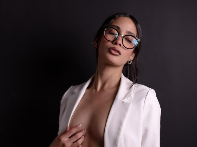 JennTylor, con los ojos cerrados, lleva un blazer blanco abierto que deja ver sus pechos. Tiene el pelo oscuro, gafas y una mano cubriendo su pezón. Fondo oscuro..