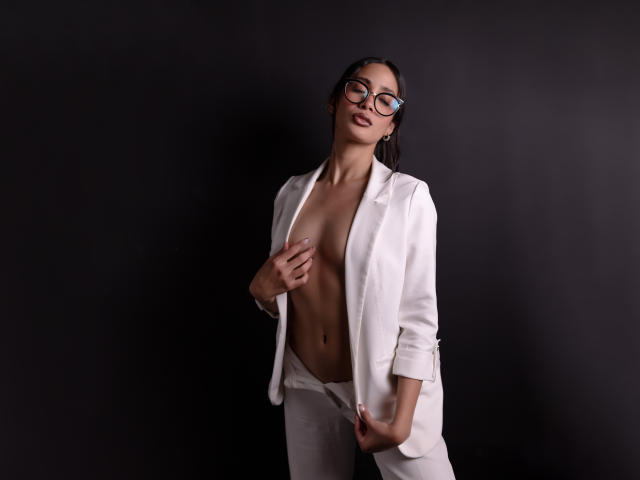 JennTylor mit Brille, trägt ein weißes, offenes Hemd, das ihre kleinen Brüste und flachen Bauch zeigt, Hände an der Hose, dunkler Hintergrund, blickt nach unten, leicht lächelnd..