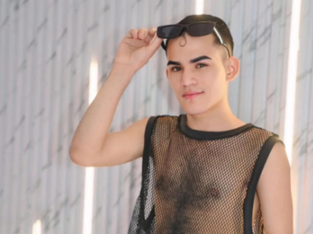 EloneMuskcle, um jovem com cabelo preto curto, vestindo uma blusa preta de malha sem mangas, com óculos de sol pretos na cabeça, mostrando um leve sorriso, parecendo fofo..