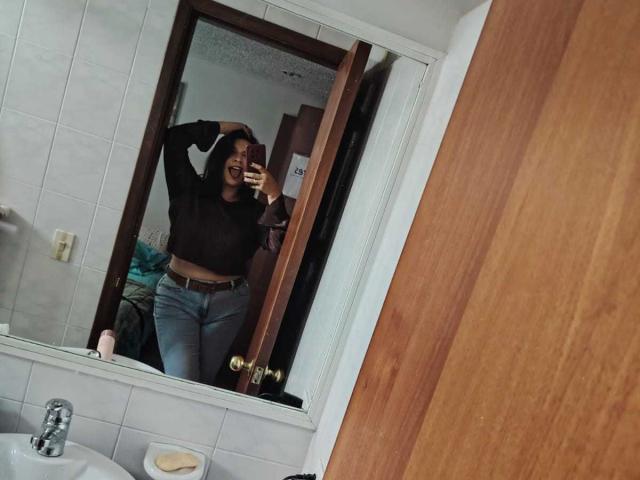 ZoeyGrey taking a mirror selfie in a bathroom, wearing a black long-sleeve top and blue jeans, with one hand on her head, showing her tits and ass.