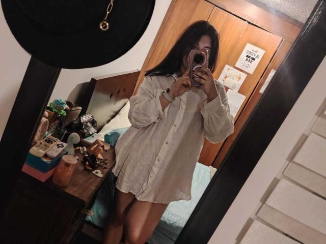 ZoeyGrey, com cabelos longos e escuros, vestindo uma camisa branca larga, em uma selfie em frente ao espelho, mostrando suas pernas e uma pequena parte das nádegas, segurando um telefone rosa..