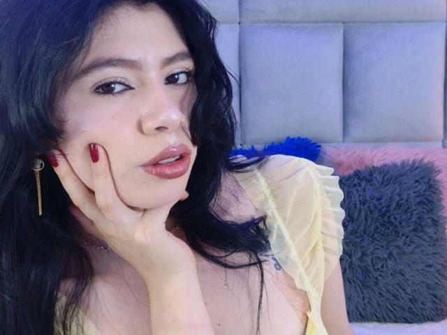 LindaMancini with dark wavy hair, red lipstick, and red nails, hand on cheek, wearing a sheer yellow top, blue earrings, and a subtle tattoo on her chest.
