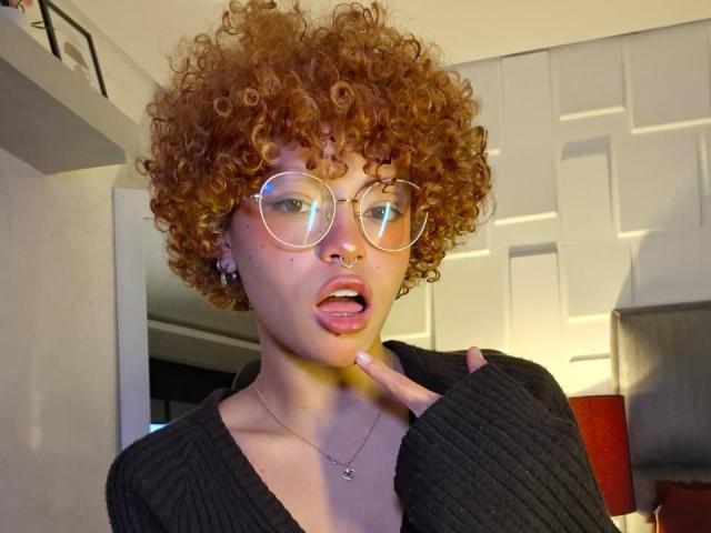 KeiryGinger with curly red hair, glasses, nose ring, and tongue out, touching her chin, wearing a black ribbed sweater, has small tits, and a necklace in a selfie.