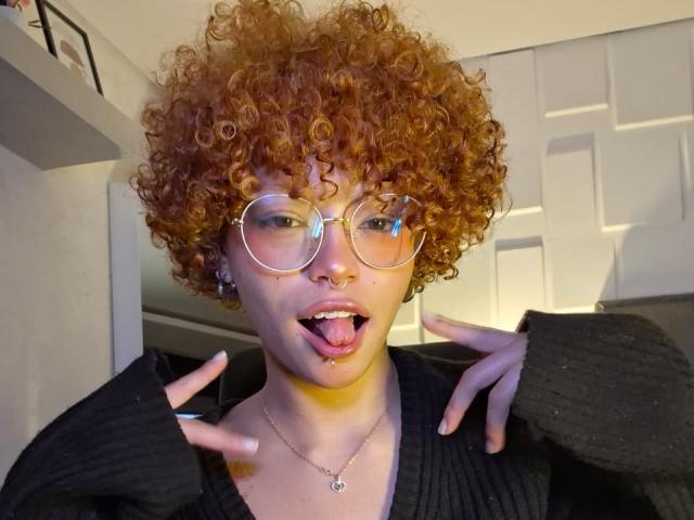 KeiryGinger with curly red hair, large round glasses, black sweater, tongue out, nose ring, and necklace, hands near face, in a well-lit room, looking playful and cute.