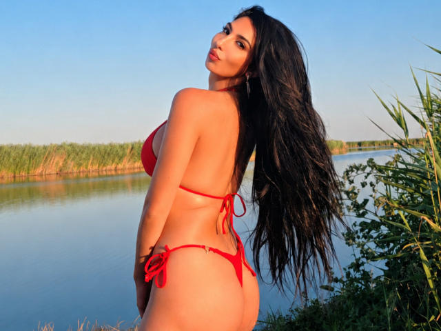 BrunetteBabe69 mit langen, schwarzen Haaren in einem roten Bikini, die ihren Rücken und Hintern zeigt, und die über ihre Schulter schaut, steht an einem ruhigen Gewässer mit grünen Schilfhalmen..