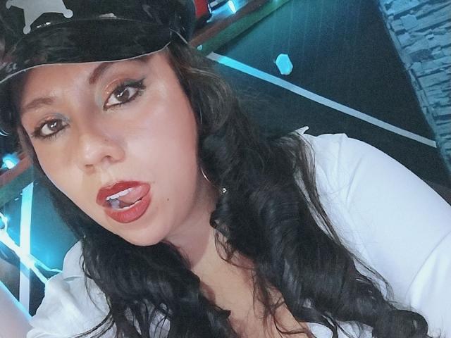 LexyDom mit schwarzen, gewellten Haaren, die ein weißes Hemd und eine schwarze Mütze trägt, mit ihrer rot gespitzten Zunge herausgestreckt, wobei ein Metall-Zungentattoo sichtbar ist, mit blauen Lichtern im Hintergrund..