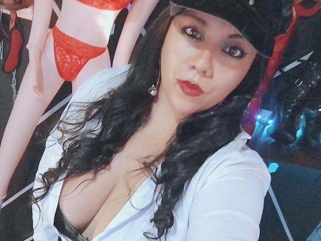 LexyDom mit lockigem, schwarzem Haar, rotem Lippenstift und einem weißen Hemd, das ihre großen Brüste zeigt, trägt eine schwarze Mütze und steht vor einer Puppe in roter Spitzendessous..