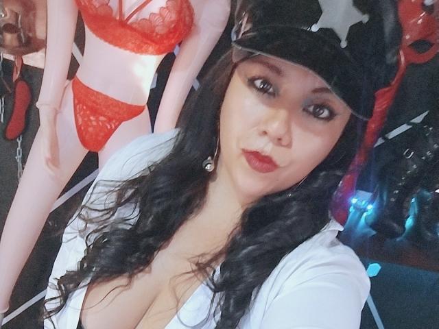 LexyDom mit langen, schwarzen Haaren, die eine schwarze Mütze und ein weißes Hemd trägt, zeigt auf einem Selfie ihre großen Brüste. Im Hintergrund ist eine rote Puppe mit großen Brüsten zu sehen..
