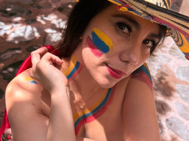 FreyaraVenwo with colorful paint on face and chest, wearing a straw hat, holding her hair, showing her tits, and a red cloth, smiling at the camera.