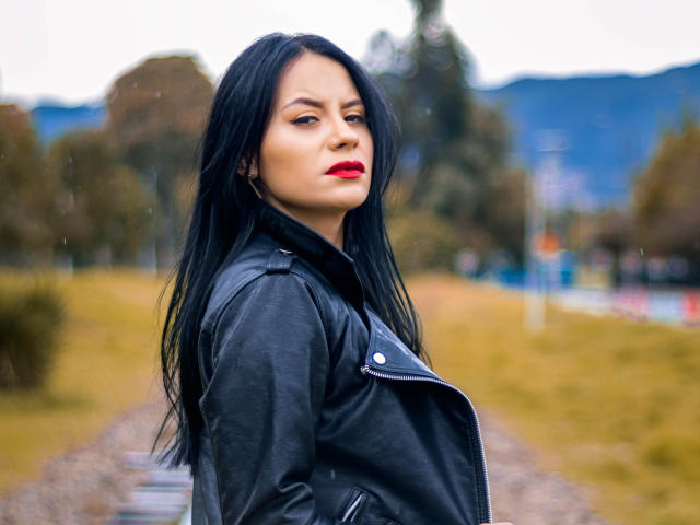LiaTayloor con giacca di pelle nera, rossetto rosso e lunghi capelli neri, che guarda oltre la spalla con un'espressione seria, all'aperto, cielo nuvoloso, in piedi su un sentiero di ghiaia..